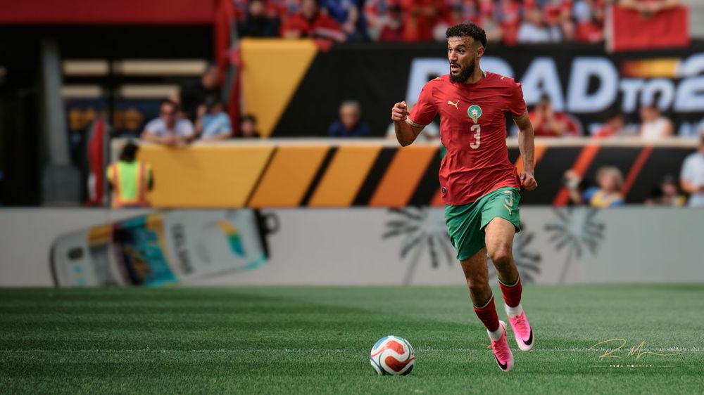 Morocco footballer in red jersey number 3 dribbles soccer ball on field during competitive match.