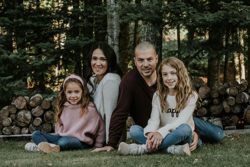A happy family of four sits together on grass in front of a log pile in a forest.