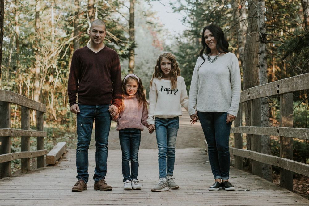 A family of four walks hand in hand on a wooden bridge surrounded by autumn foliage.