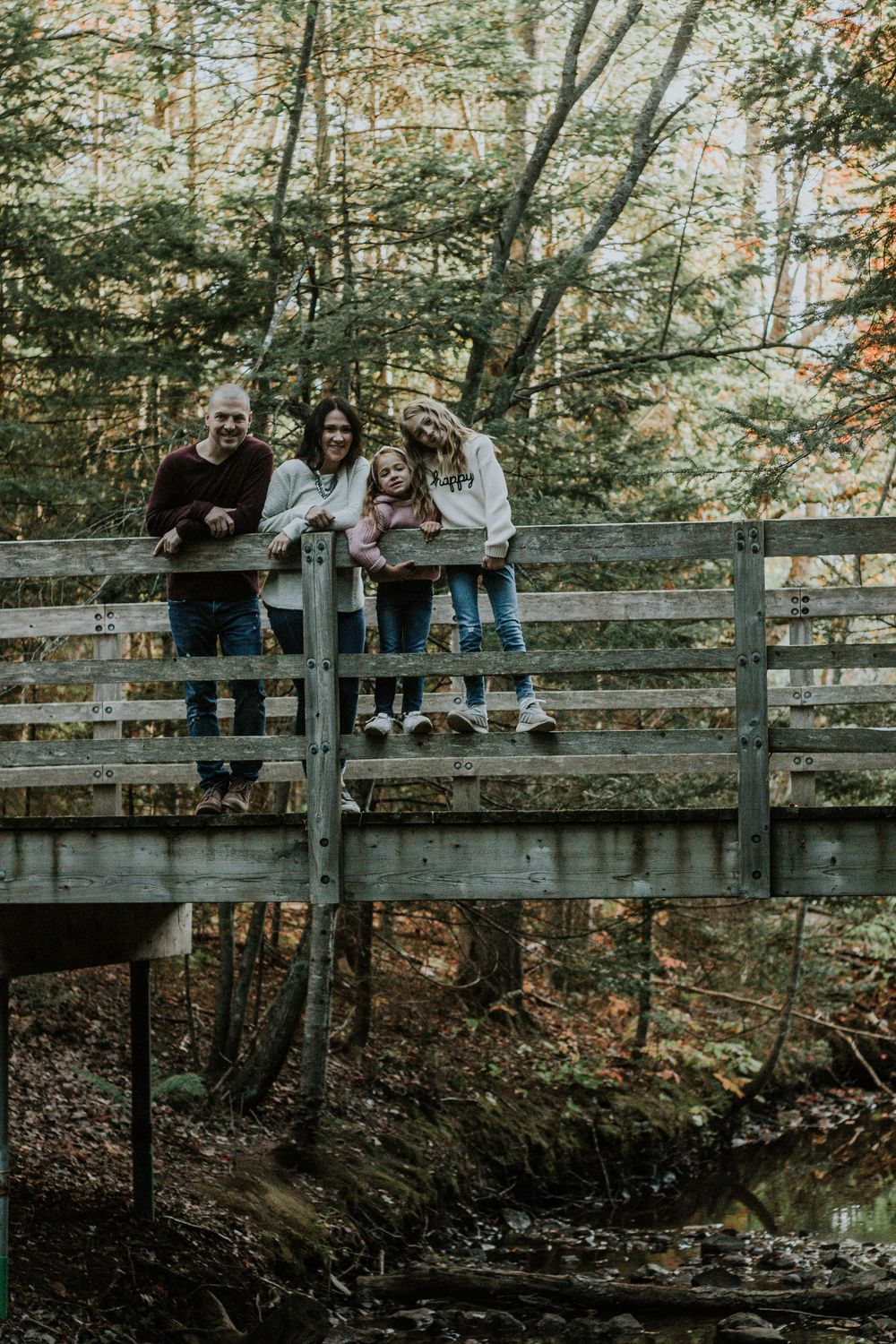 A family of four smiles together while standing on a rustic wooden bridge in autumn woods.
