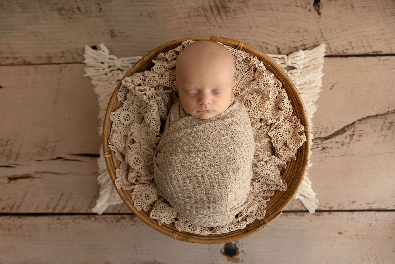 A swaddled infant rests in a wooden bowl lined with lacy fabric on a rustic white wooden floor.