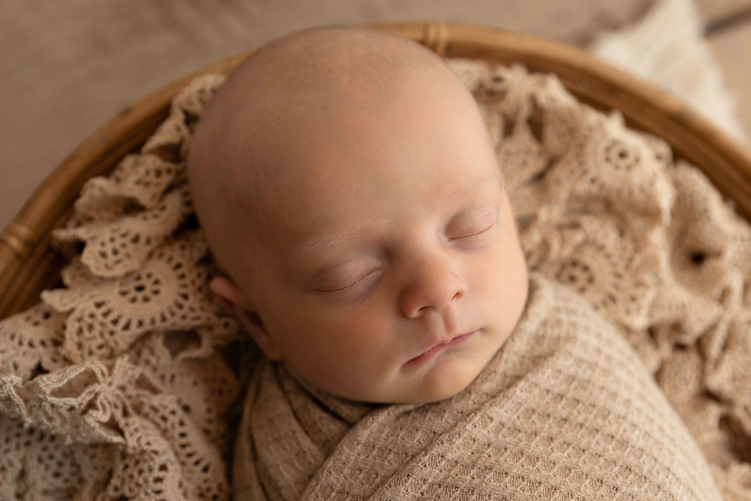 Sleeping newborn wrapped in beige knitted blanket on tan lace fabric in natural lighting.