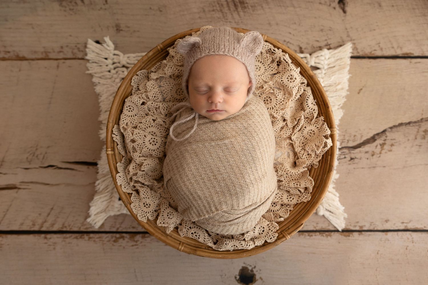 Newborn baby swaddled in beige wrap lying in round wooden bowl with lace on white wooden floor.