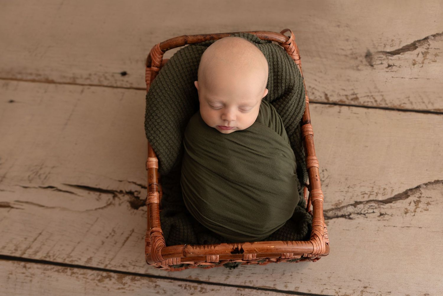 A small figure wrapped in a dark green blanket rests peacefully in a rustic wooden basket on a wooden floor.