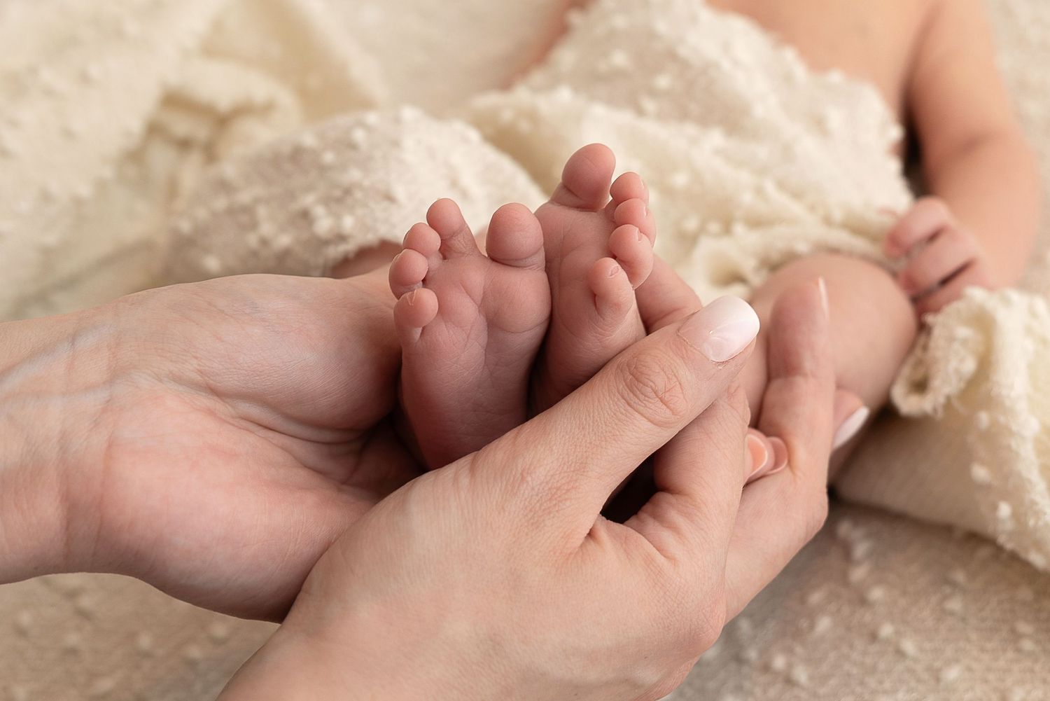 Adult hands gently cradle tiny newborn baby feet against soft beige blanket in tender moment.