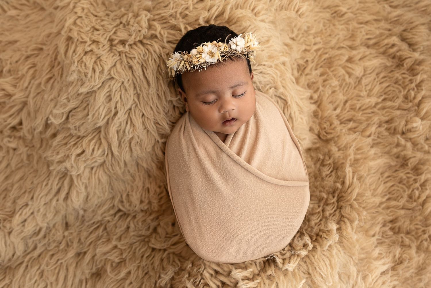 Peaceful newborn sleeping on cozy beige fur while wrapped in soft tan swaddle and wearing delicate floral crown.
