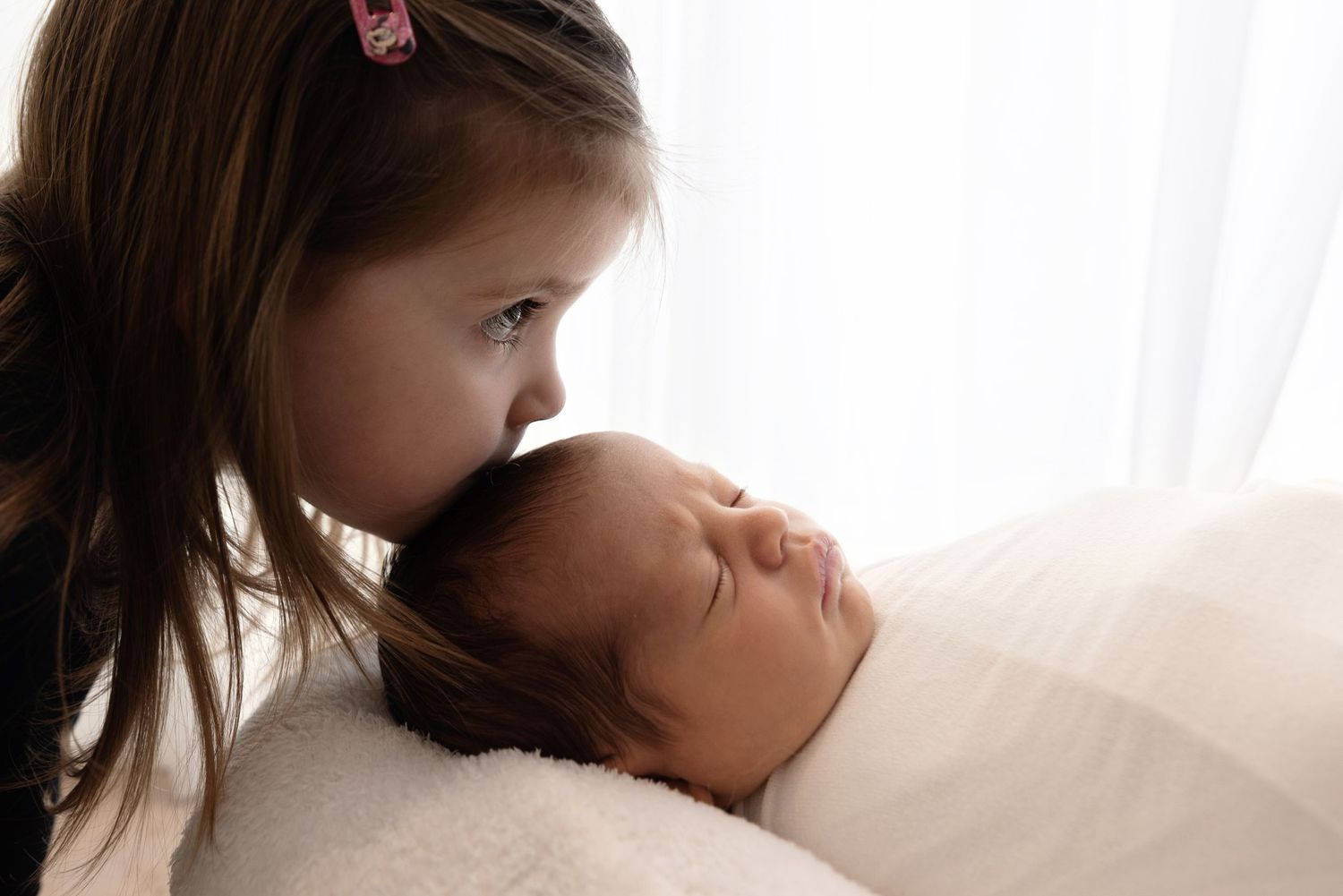A young child leans over to give a tender kiss on the forehead to a sleeping baby in a sunlit room.