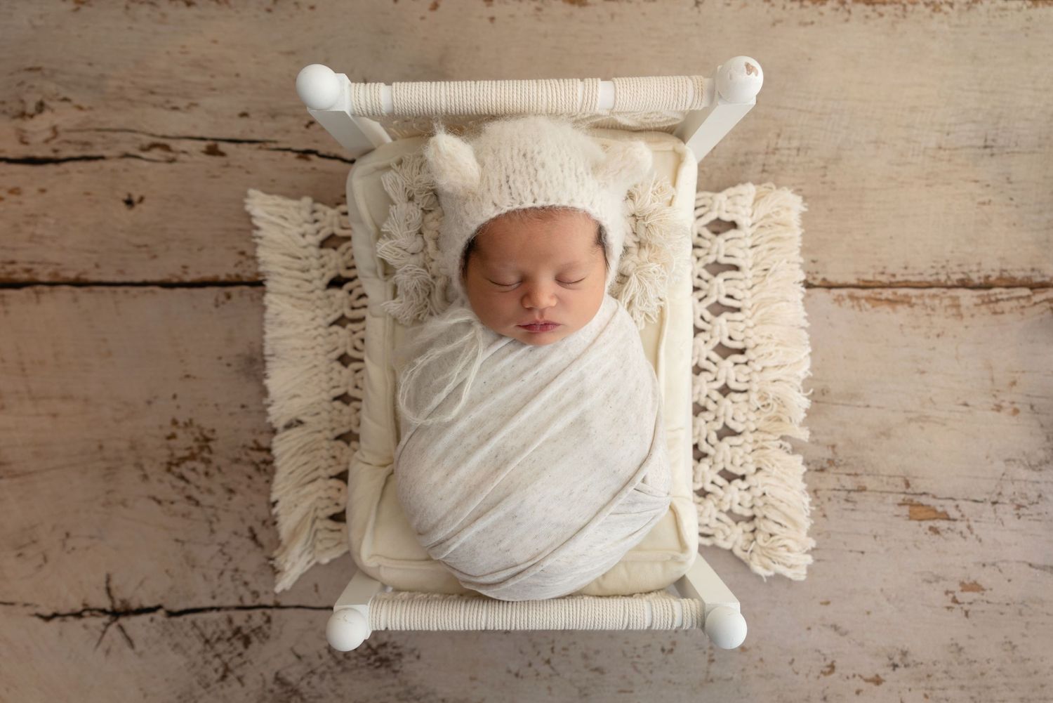 A newborn swaddled in white fabric lies on a cream macrame backdrop with rustic wooden floor beneath.