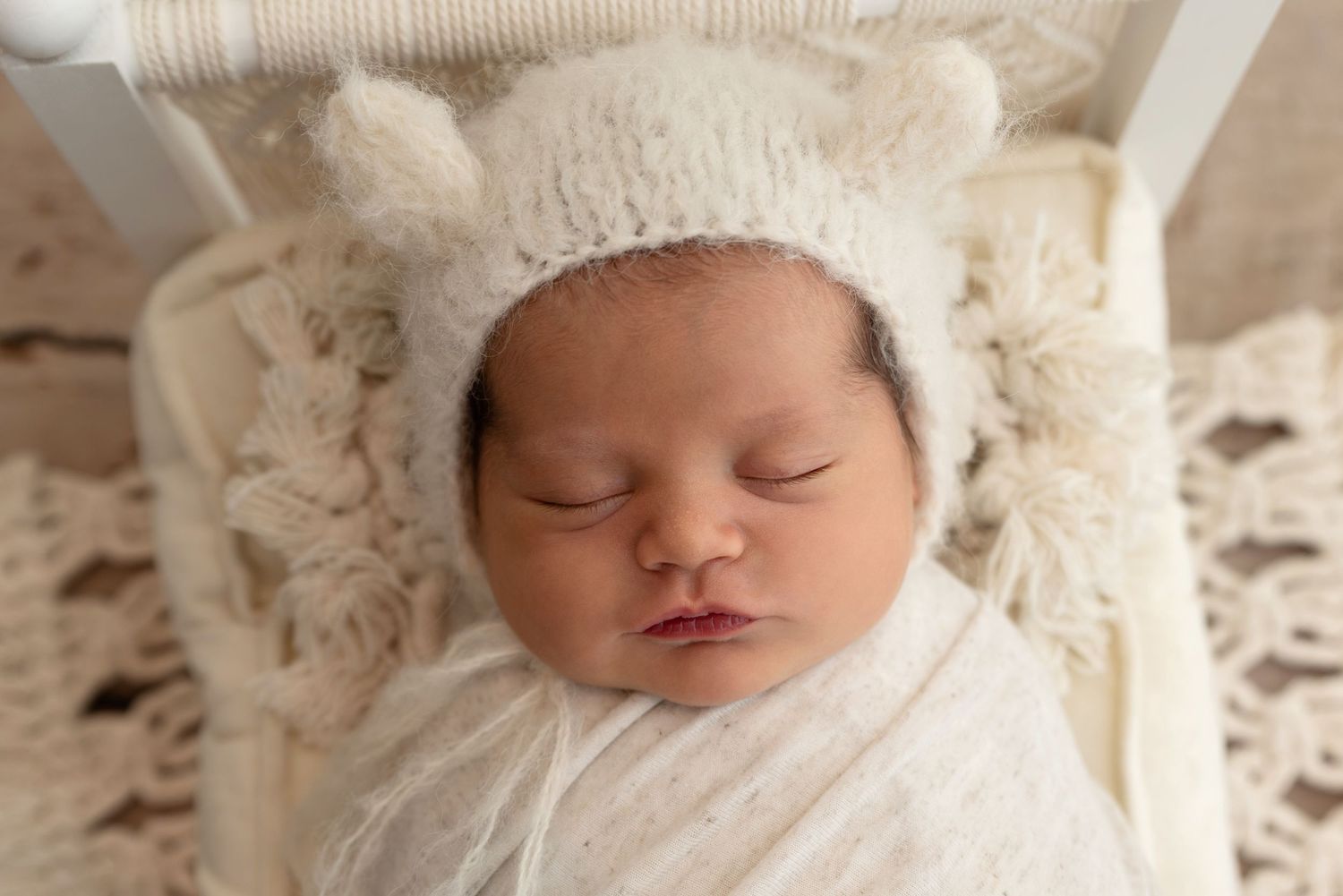 A sleeping newborn wearing a white knit bear hat is wrapped in a cozy white blanket.