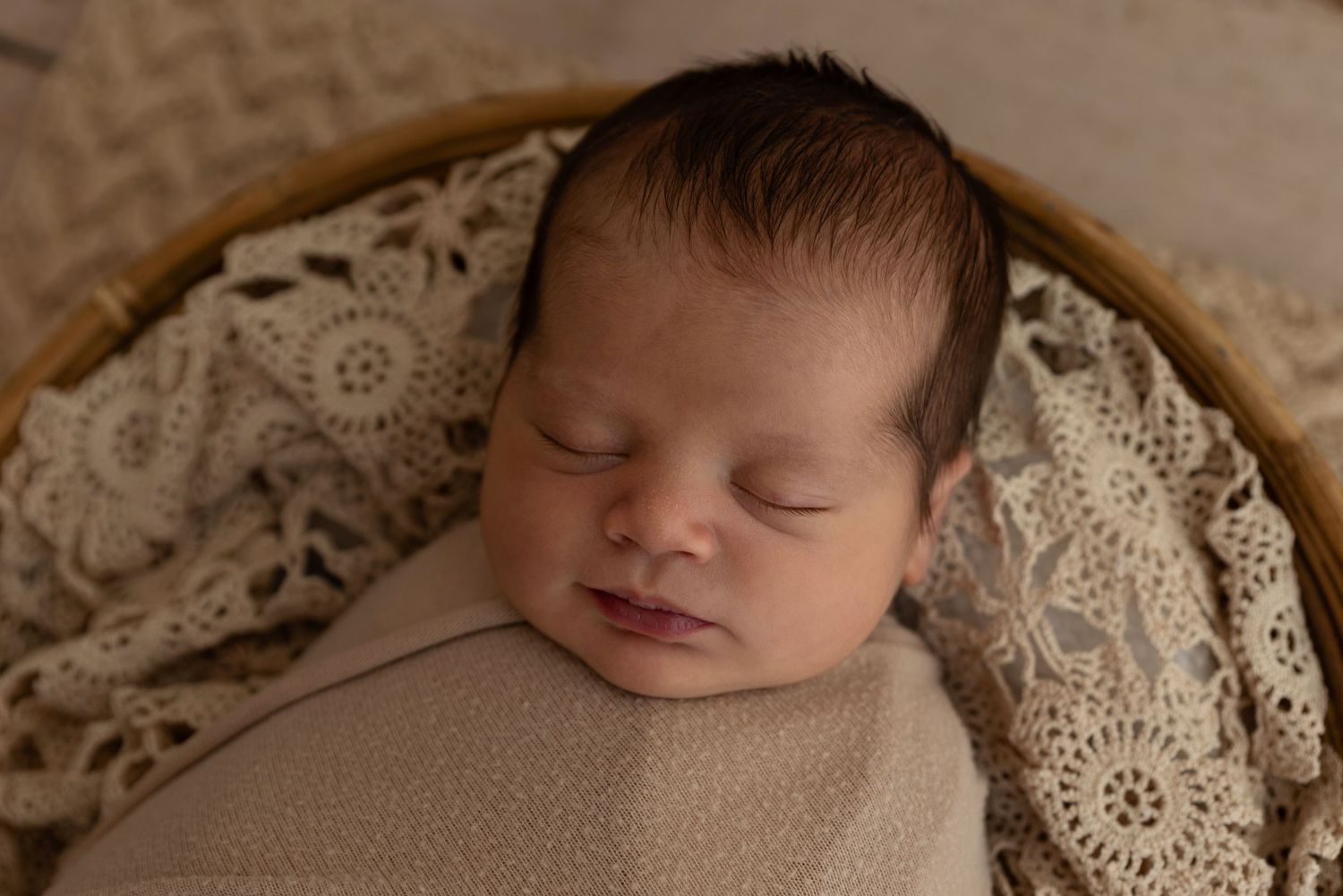 Peaceful newborn sleeps peacefully wrapped in beige fabric against lace pattern backdrop during professional photo session.