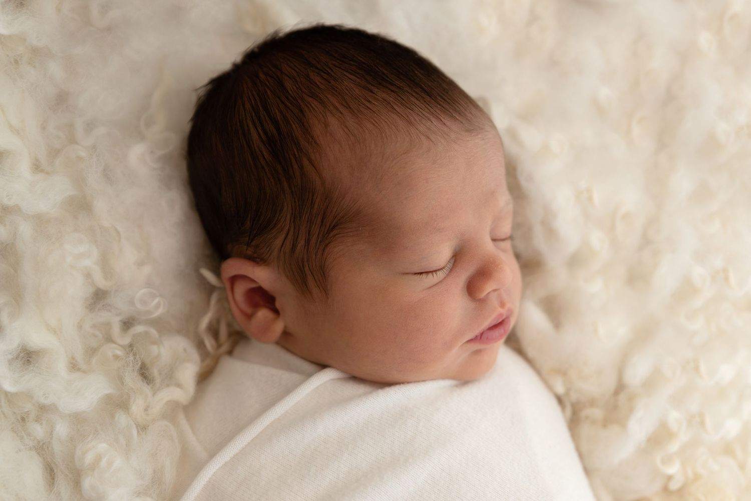 A peaceful newborn baby sleeps swaddled in white fabric against a cream-colored textured background.