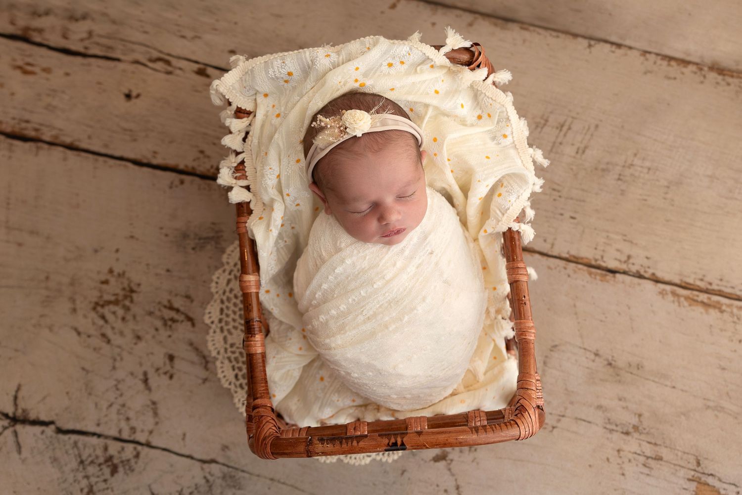 A peaceful sleeping newborn wrapped in white lace swaddle rests in a rustic wooden basket on a light wood floor.