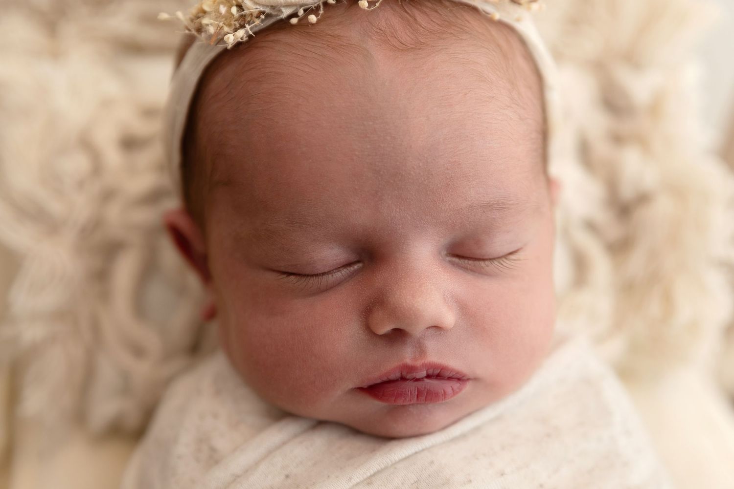 Sleeping newborn baby wearing delicate gold crown tiara rests peacefully on cream-colored fabric.