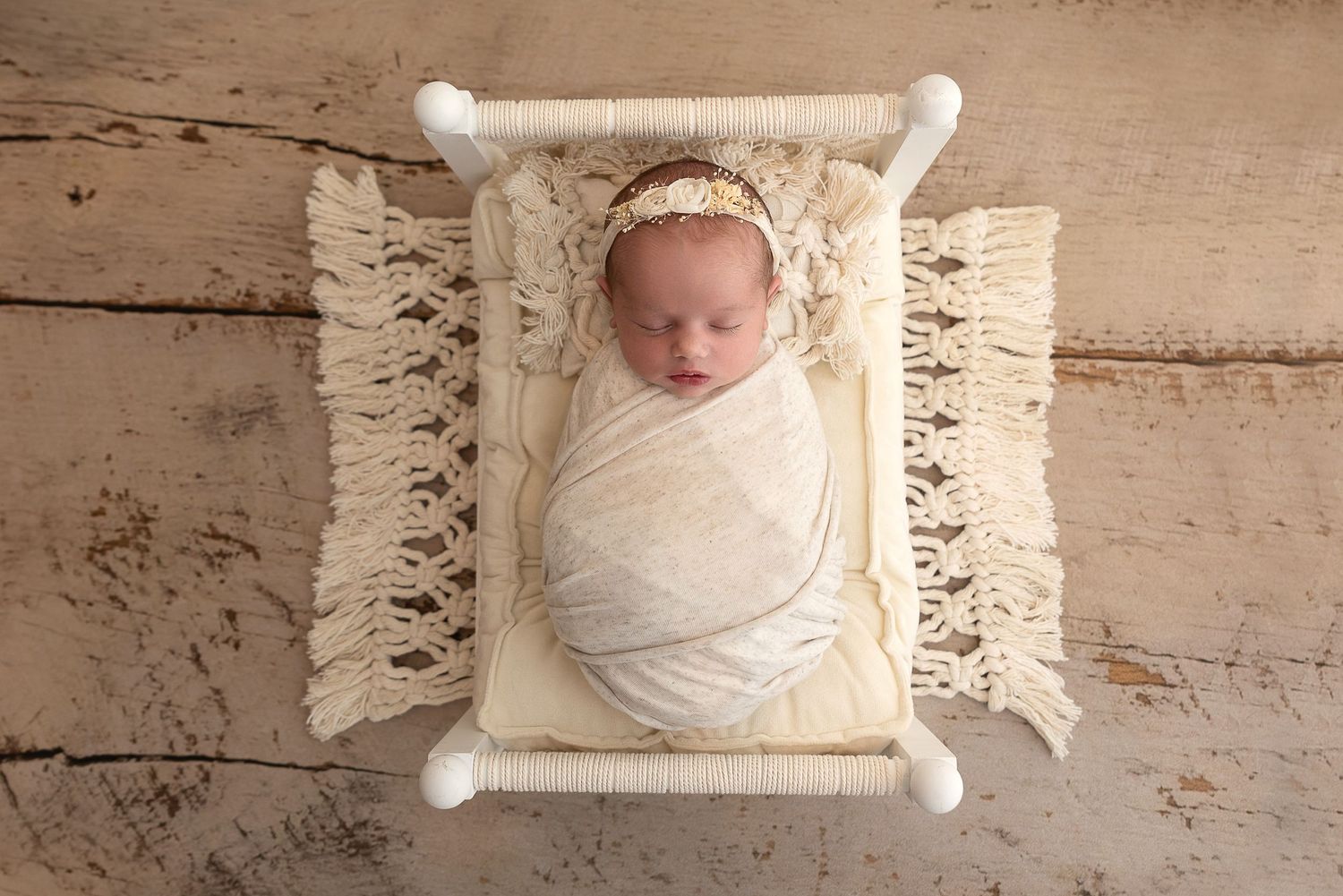 A newborn wrapped in white swaddle lying on a cream macrame bed prop with wooden frame.