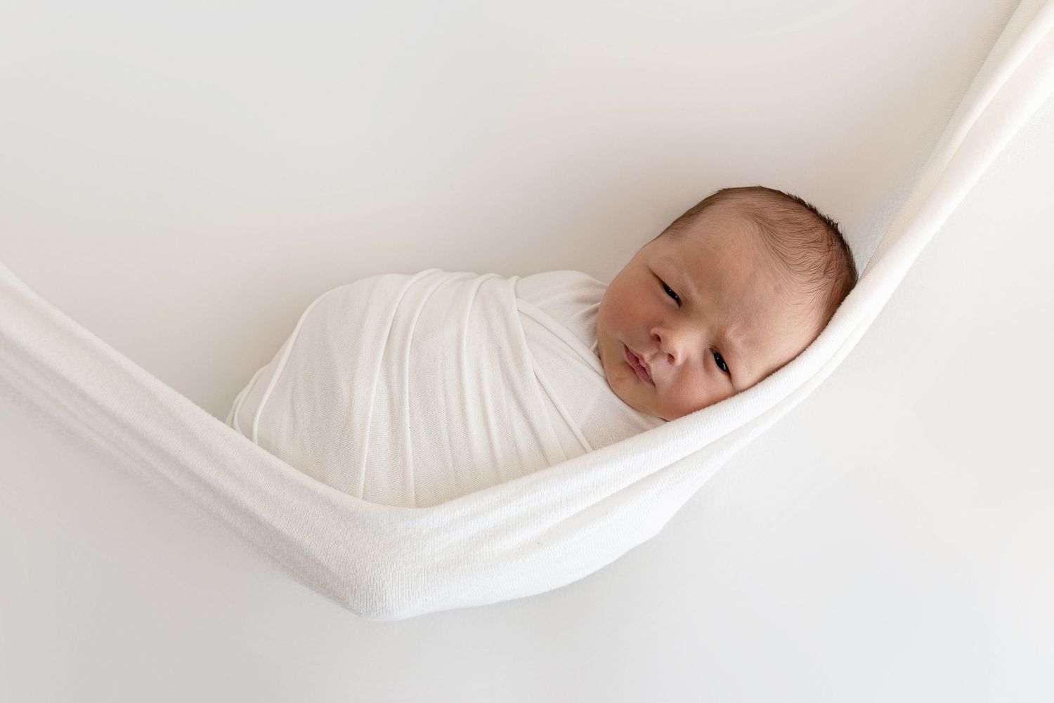 A sleeping baby wrapped in white fabric nestled in a hammock-style sling against a light background.