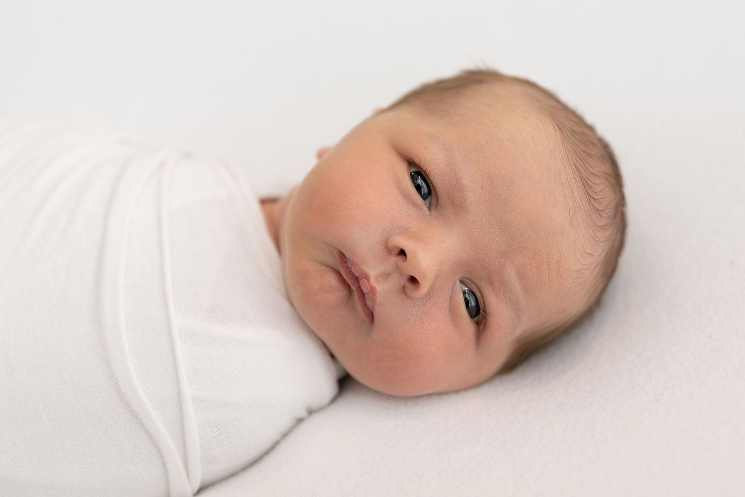 A tiny baby wrapped in soft white swaddling cloth rests on a light background.