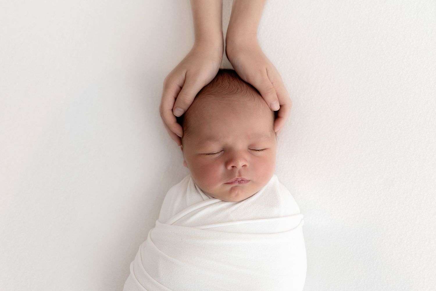 A sleeping newborn baby wrapped in white fabric rests peacefully while gentle hands cradle their head.