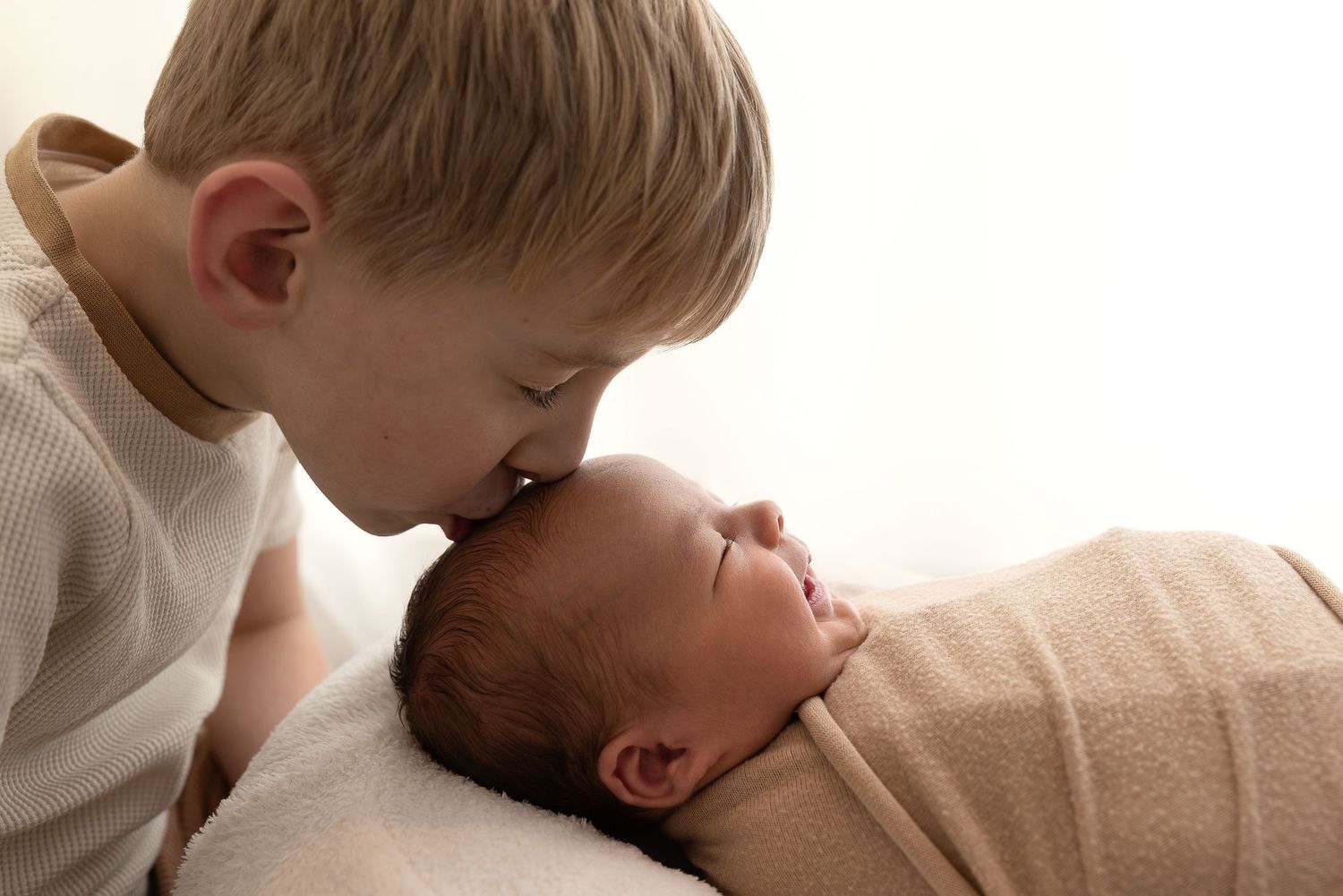 A tender moment as a young child gently kisses a newborn baby swaddled in soft fabric.