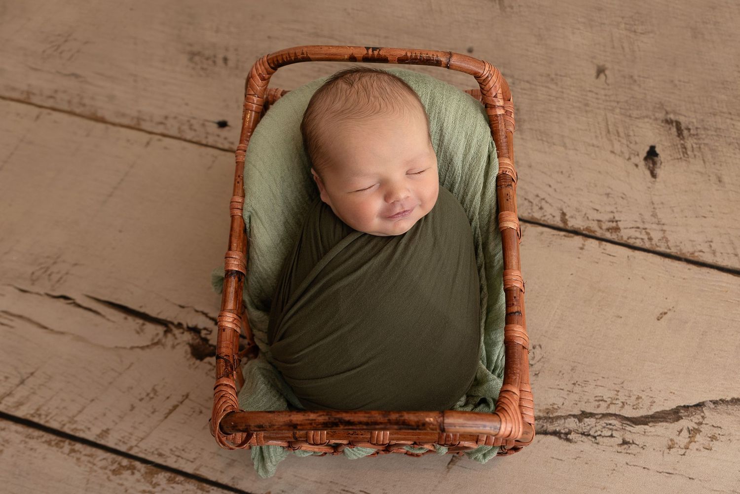Peaceful sleeping newborn swaddled in green wrap rests in small rustic wicker basket on wooden floor.