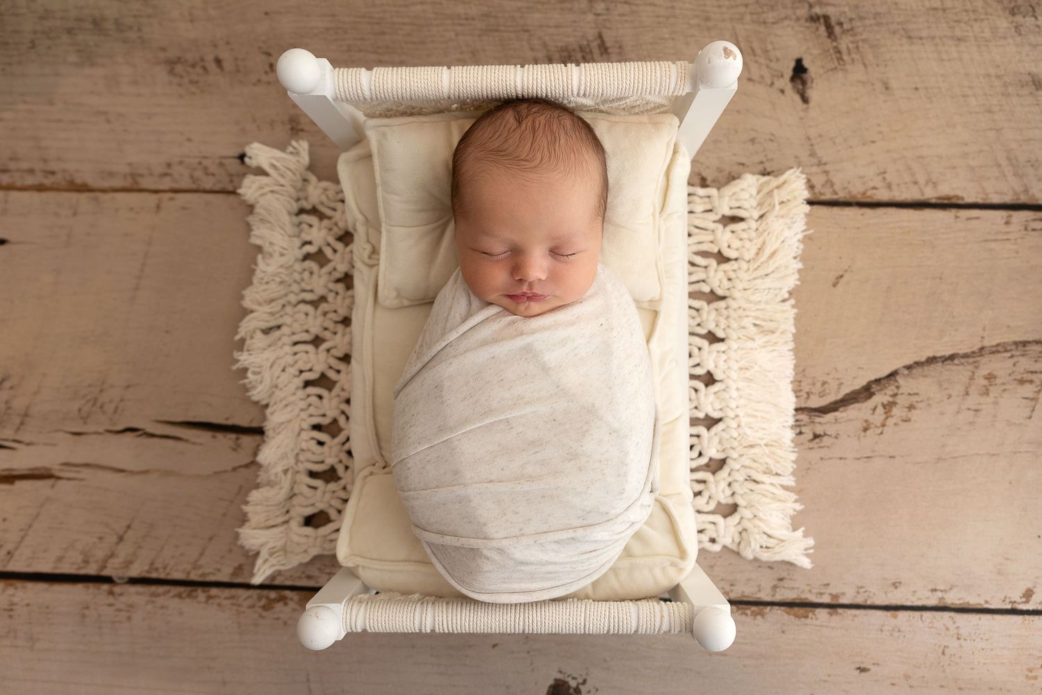 Newborn wrapped in white swaddle blanket sleeping peacefully on cream macrame cushion against wooden floor background.