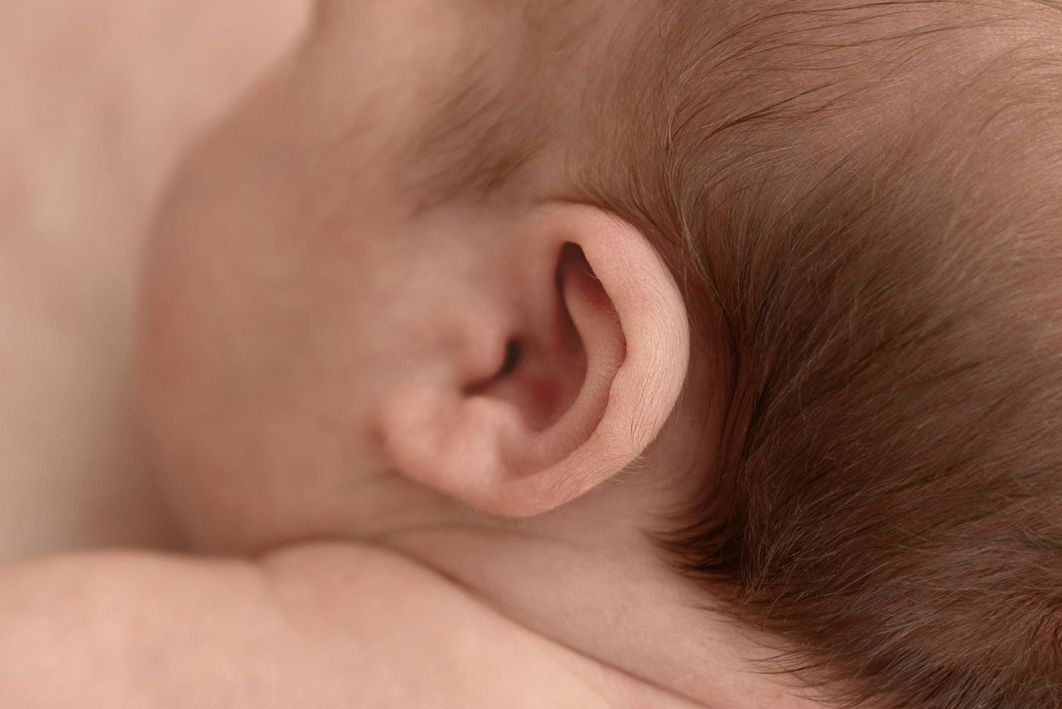 Close-up macro photograph of a baby's ear and brown hair against soft pink skin.
