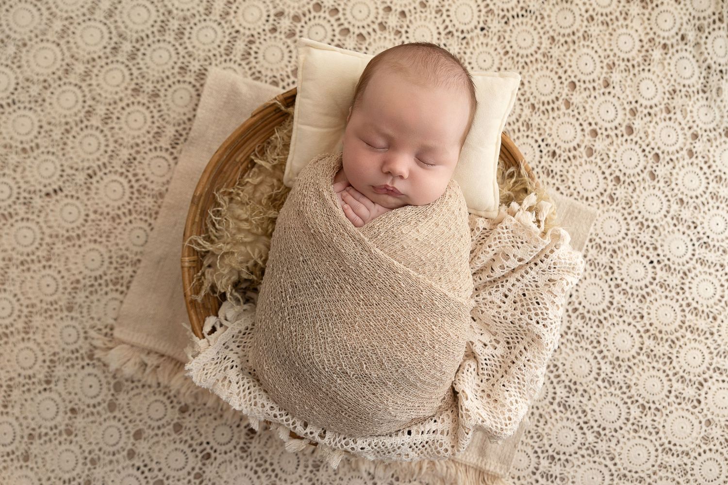 Sleeping newborn wrapped in beige swaddle blanket on textured neutral backdrop during professional photoshoot.