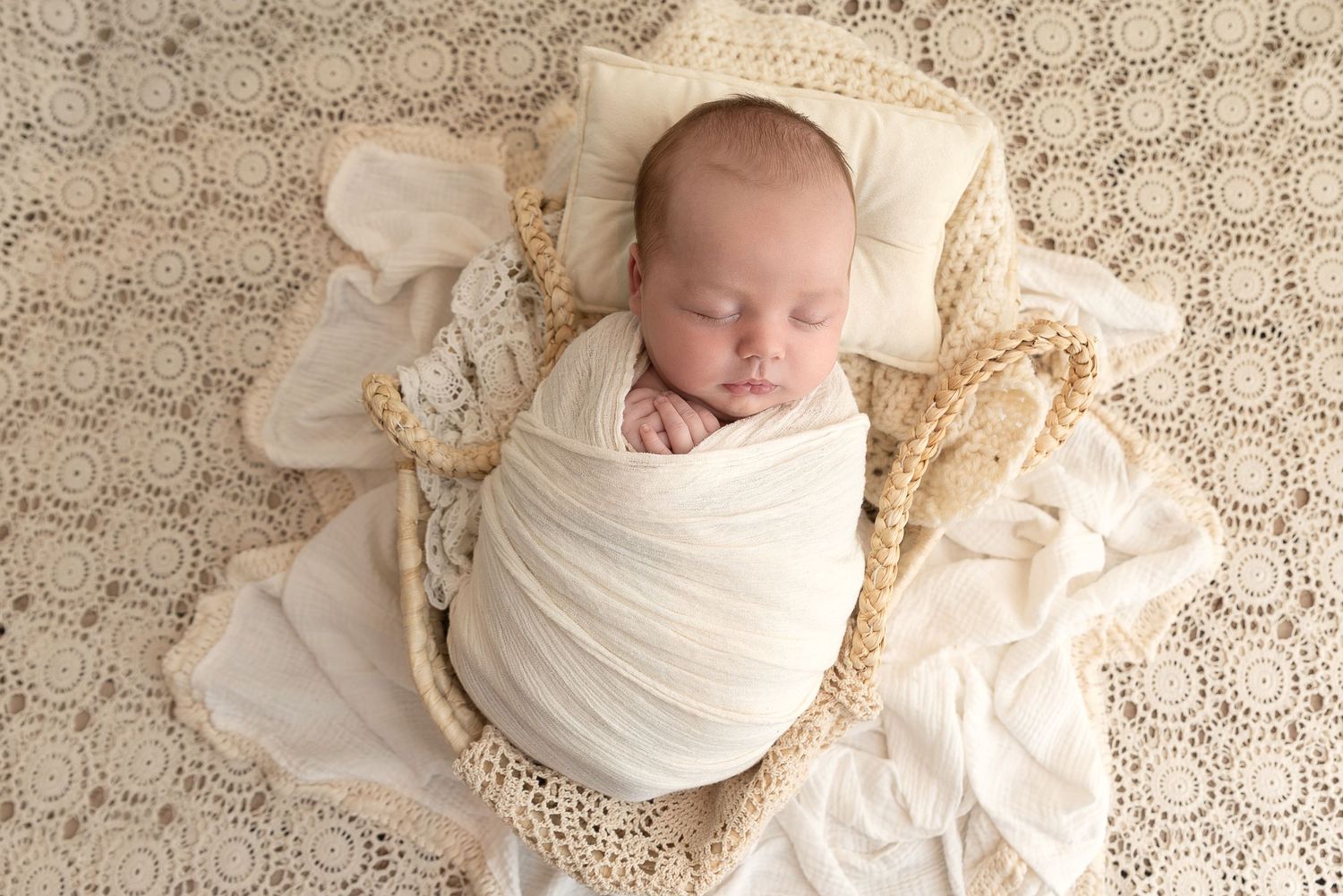 Sleeping newborn baby wrapped in white fabric lying on a textured cream-colored blanket.