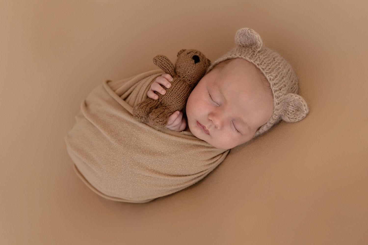 A sleeping newborn wrapped in a soft beige swaddle wearing a knitted bear hat on a neutral brown background.