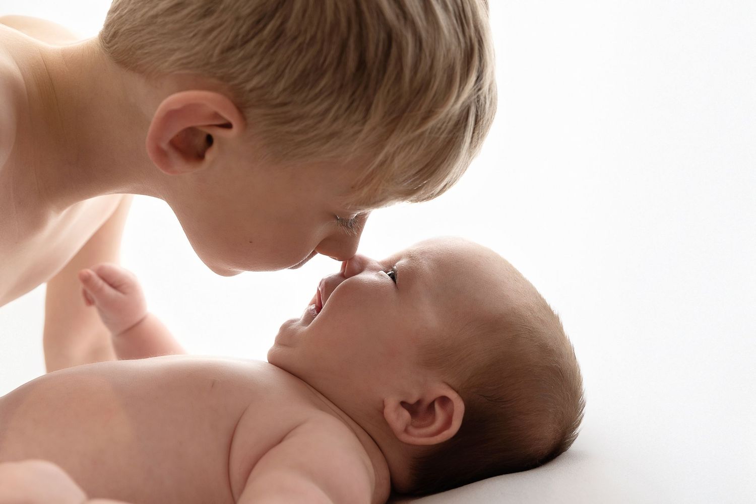 A small child leans over a smiling baby lying on a white surface in a tender moment.