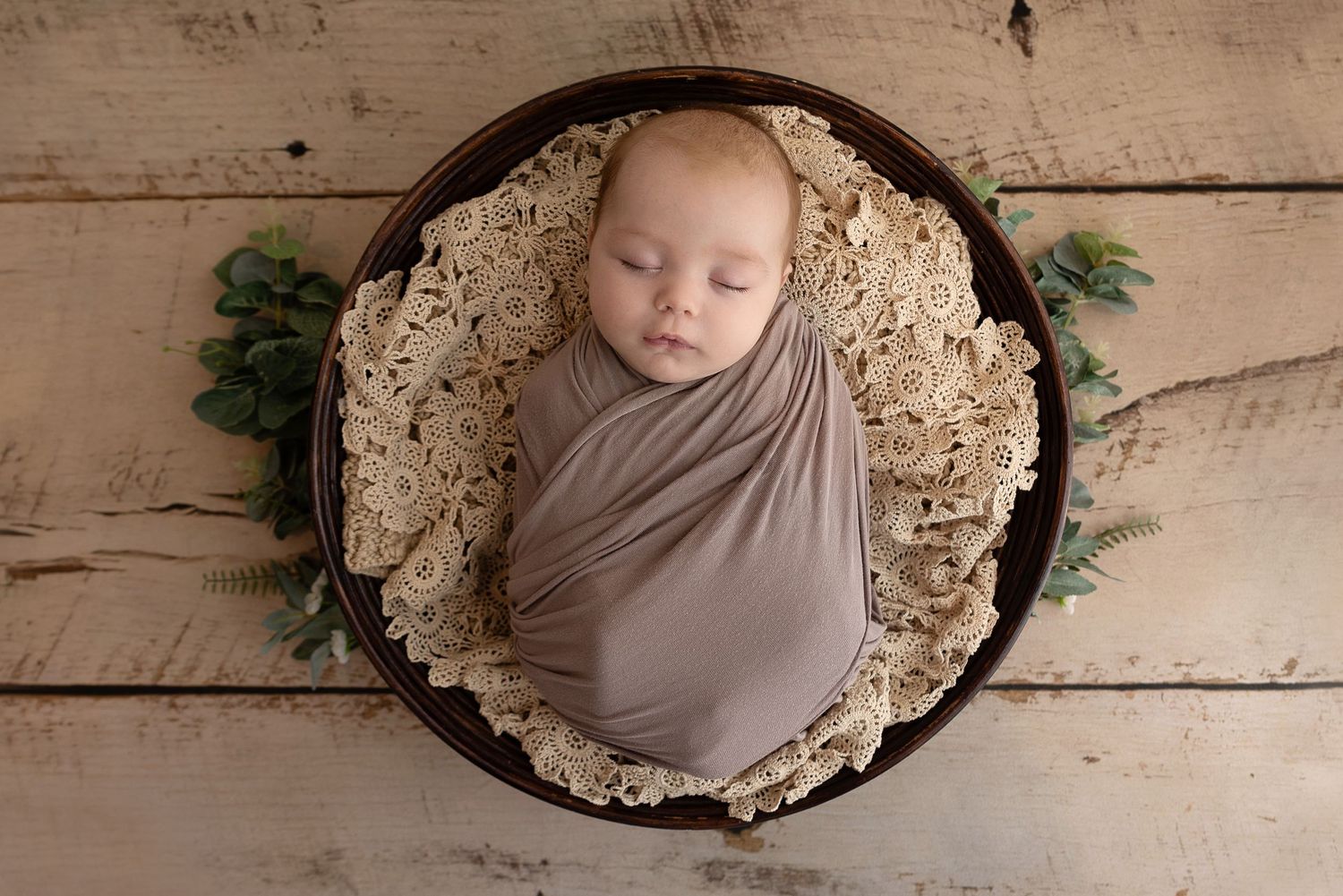 Newborn baby wrapped in gray cloth sleeping in round wooden bowl with lace cushioning and pine sprigs on wooden floor.