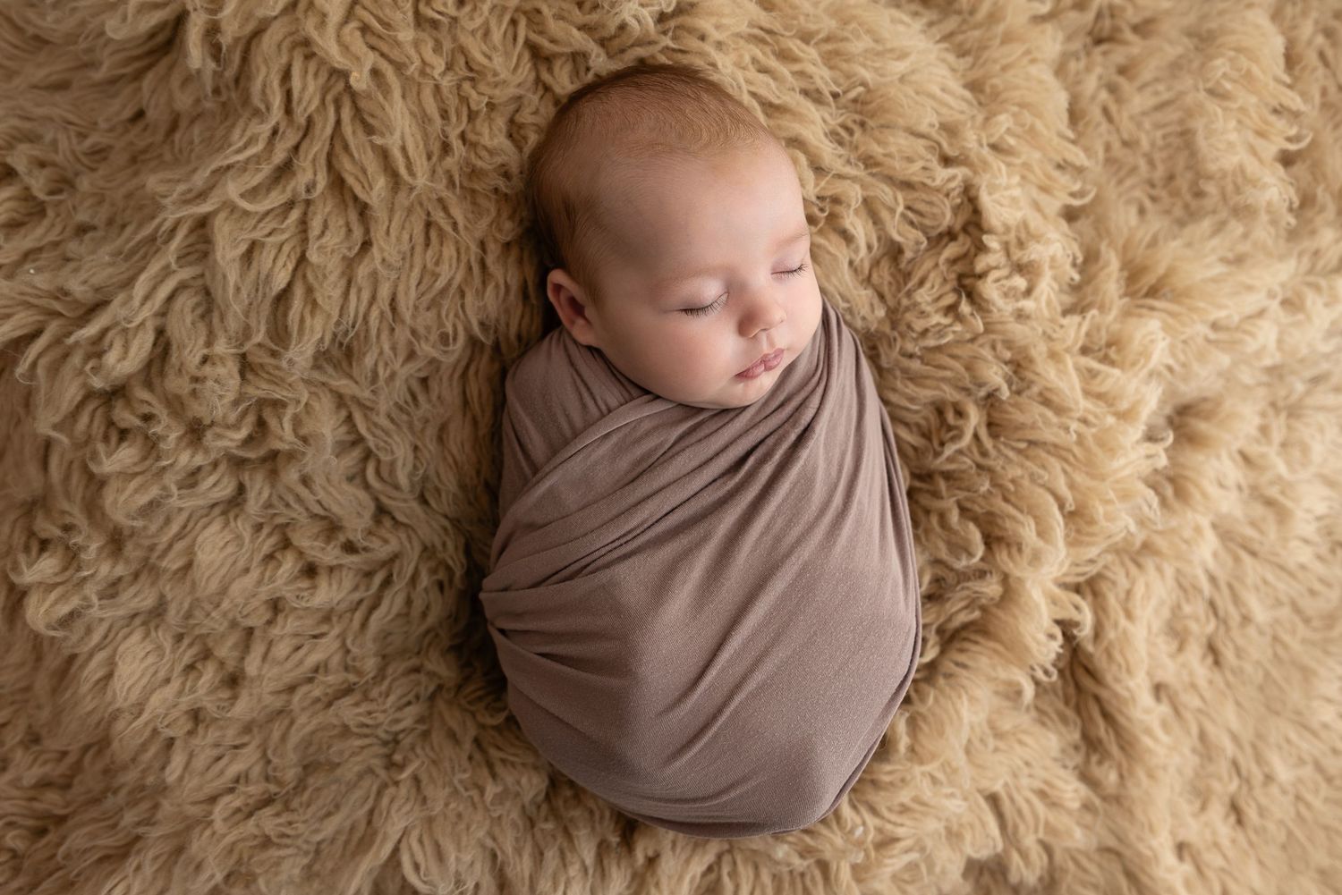 Sleeping newborn wrapped in soft brown fabric lying on fluffy beige textured background.