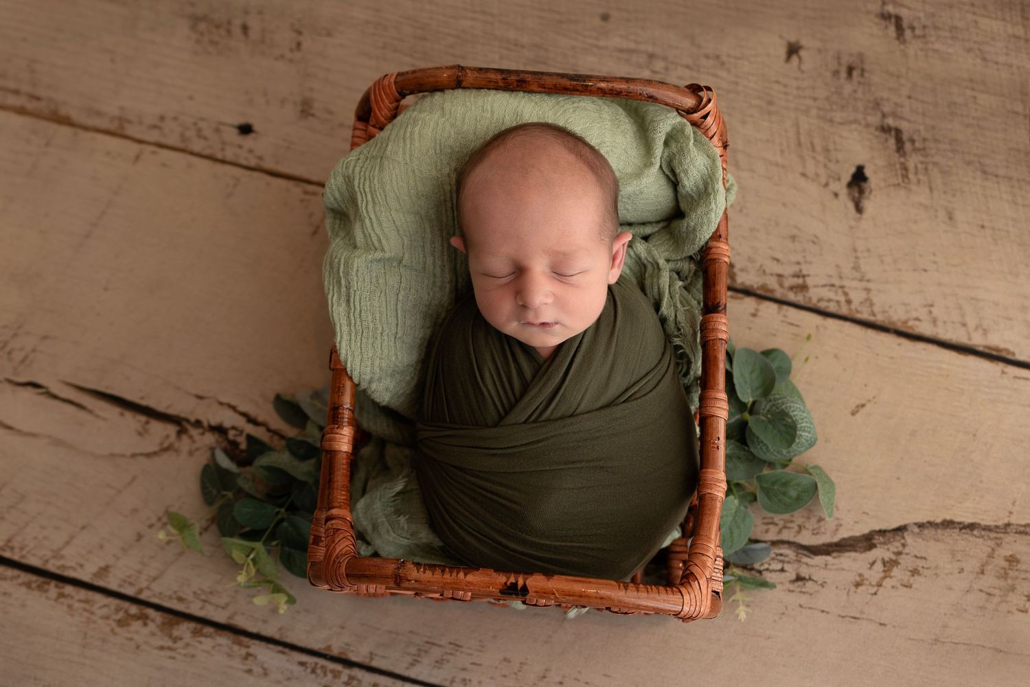 Newborn baby sleeping peacefully in wooden frame prop on rustic floor wrapped in soft green swaddle.