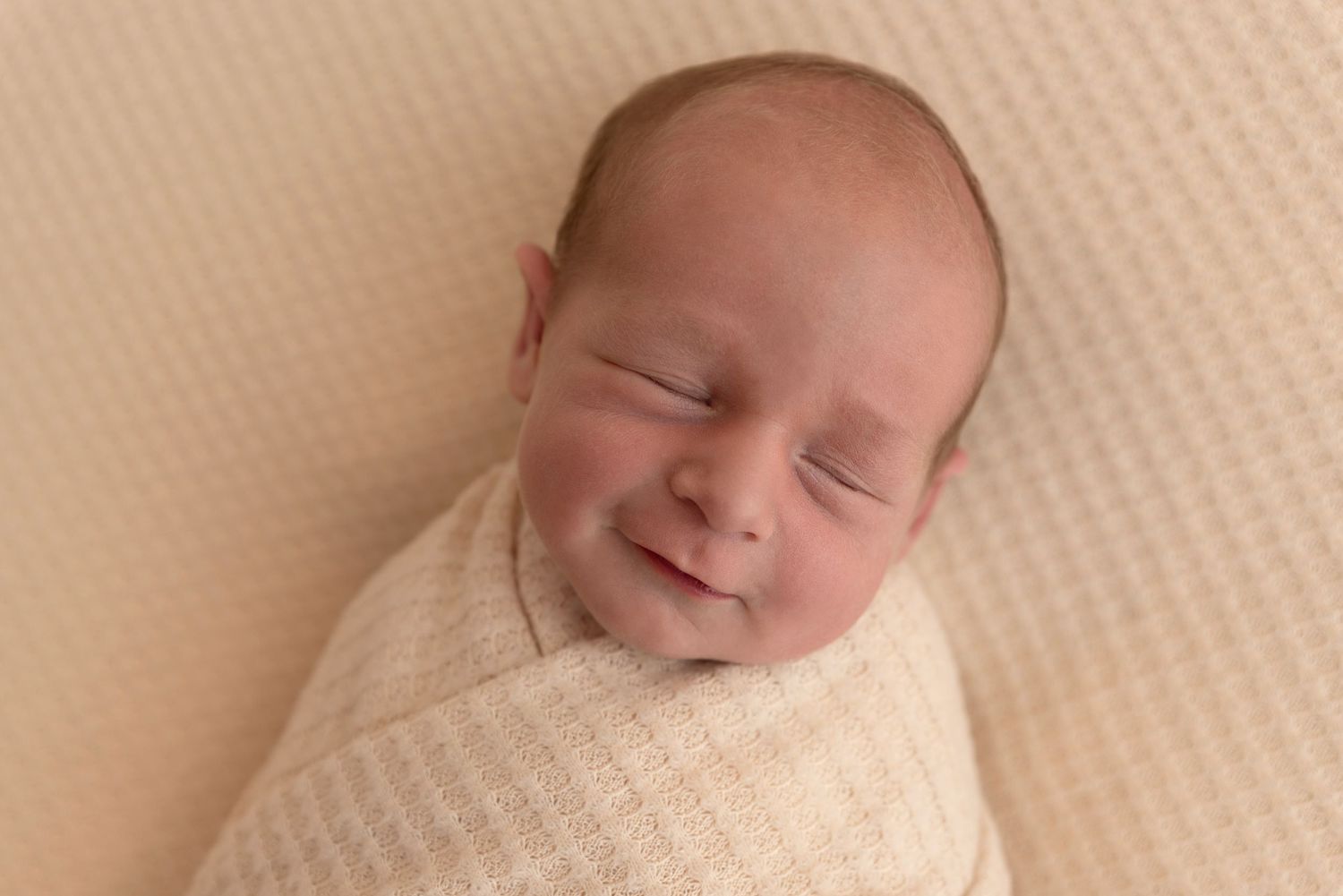 Newborn baby peacefully sleeping with a slight smile while wrapped in a soft cream-colored swaddle blanket.