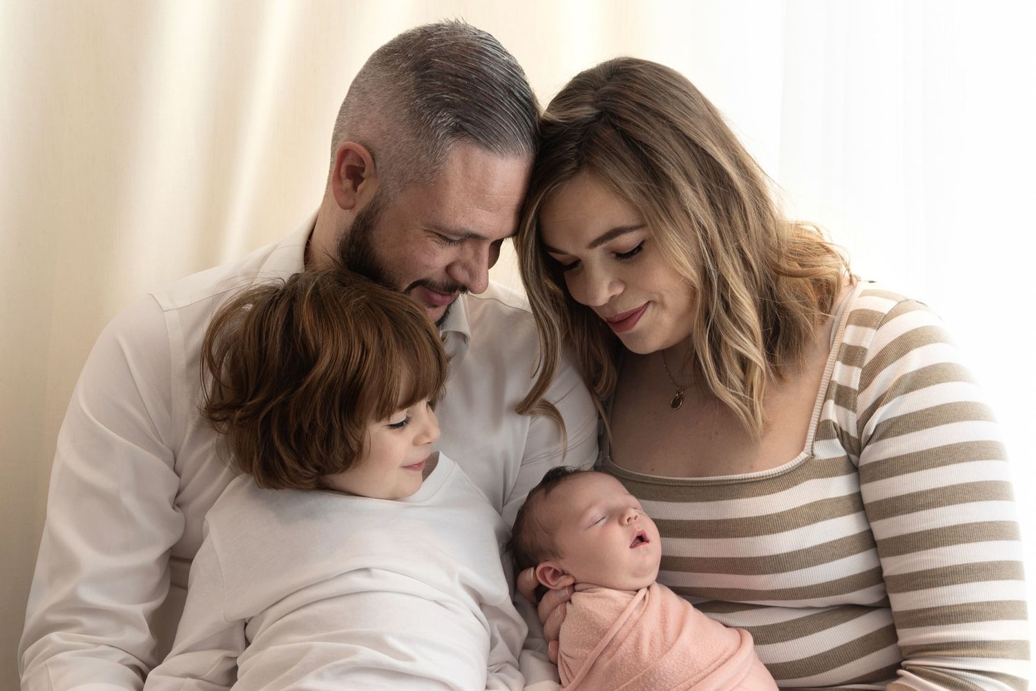 Family cuddles together in tender moment with young child and newborn baby in warm natural light.