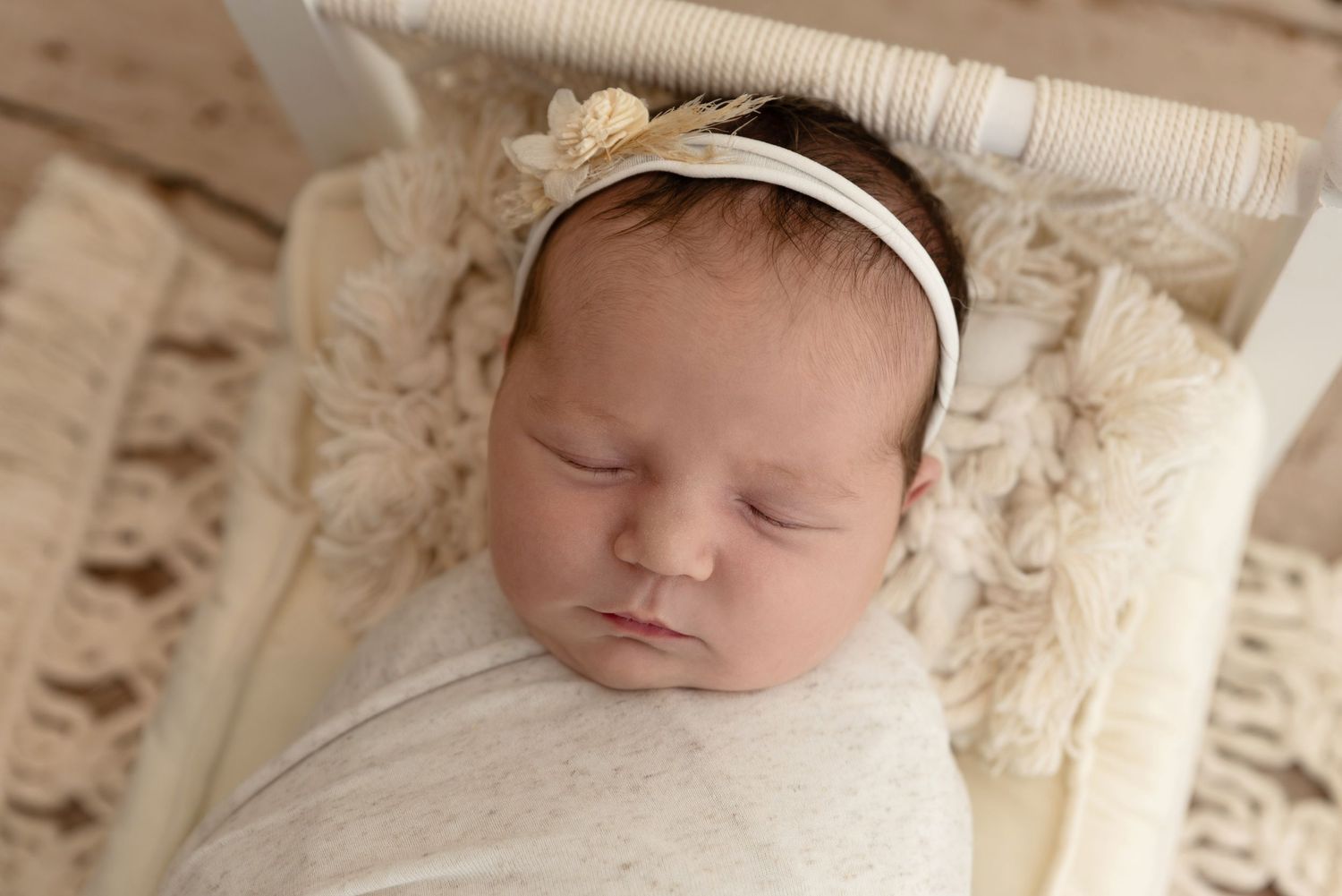 A sleeping newborn baby wrapped in white fabric with a headband rests peacefully in a cream-colored box.