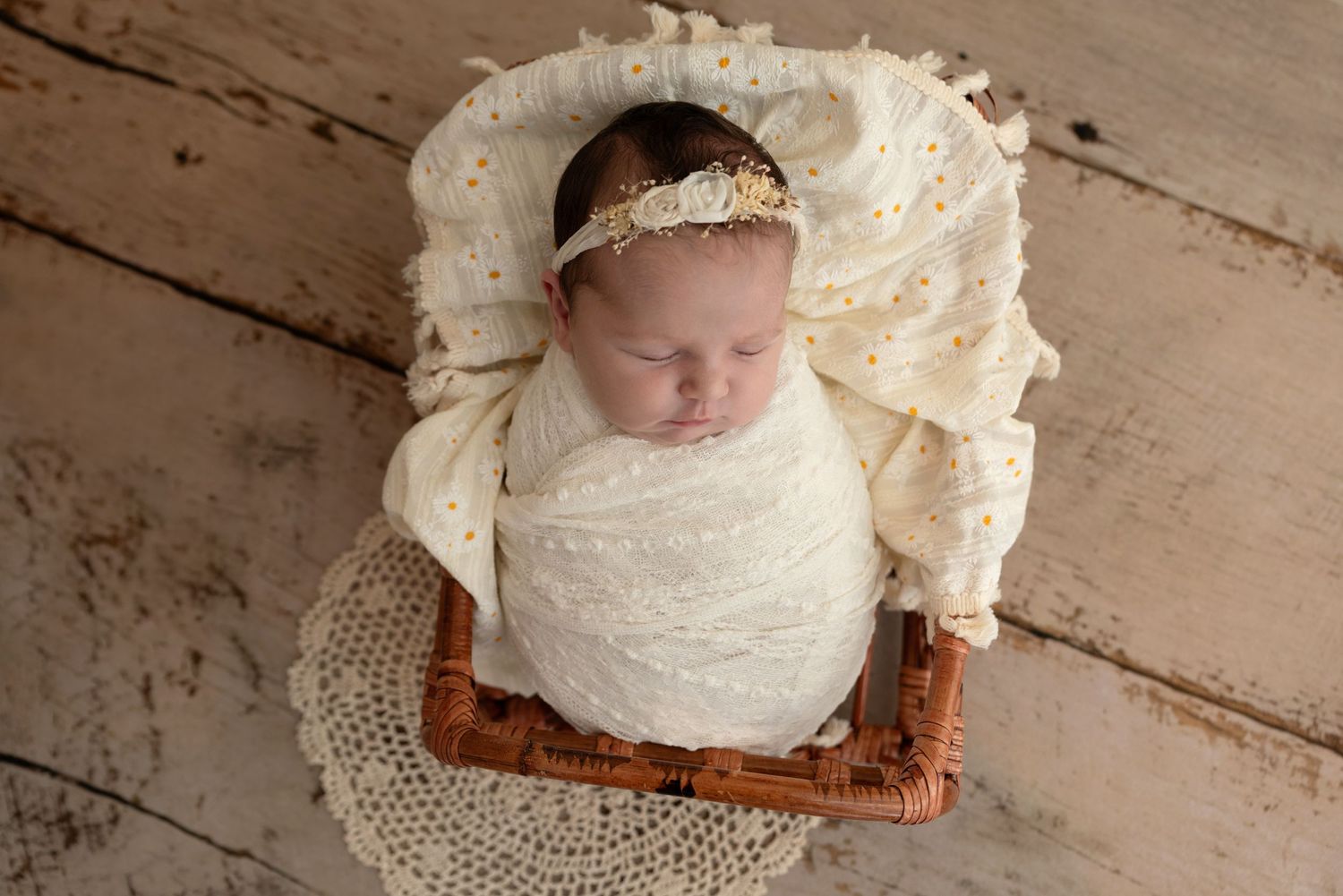 A sleeping newborn swaddled in cream fabric lies on a woven mat with rustic wooden decor.