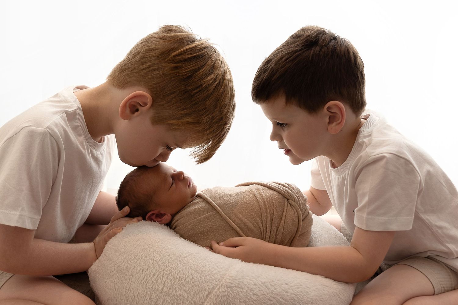 Two children in white shirts look lovingly at a newborn baby resting on a beige pillow.