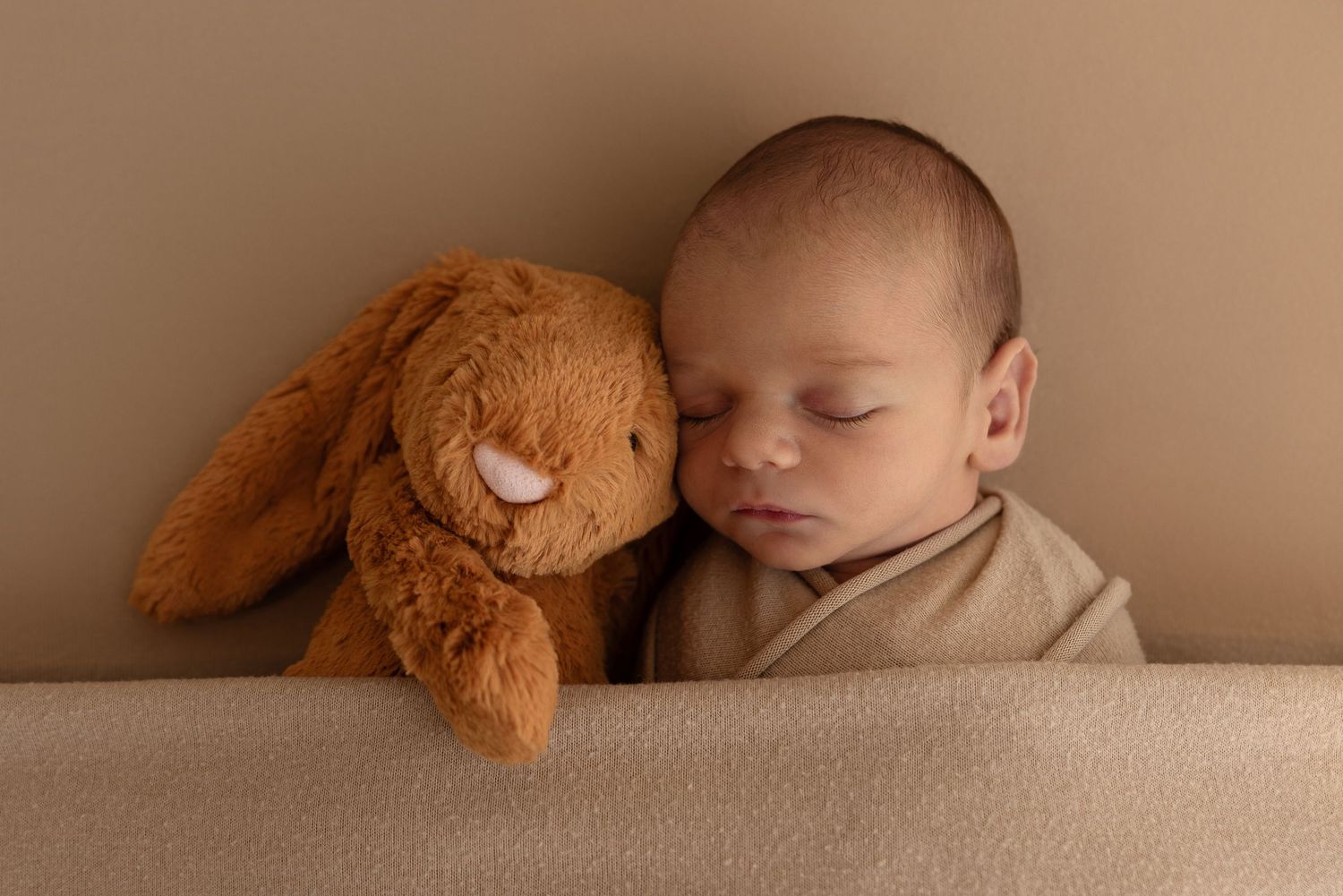 Peaceful newborn sleeps sweetly next to soft brown plush bunny on beige backdrop.