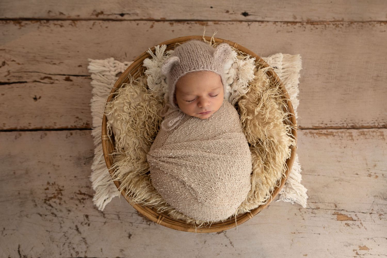 Sleeping newborn wrapped in beige swaddle lying in rustic basket with cream fringe on wooden floor.