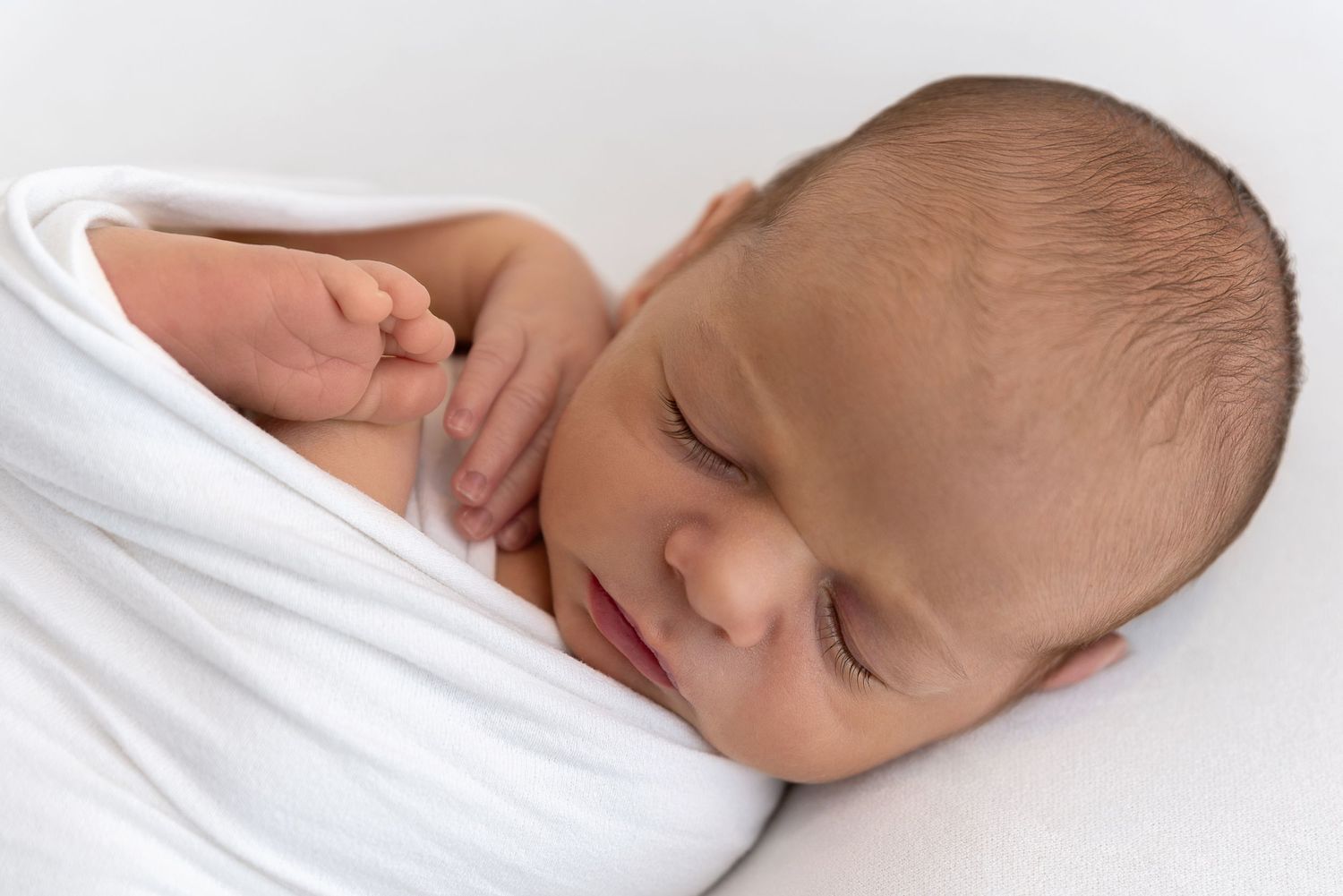 Peaceful newborn baby wrapped in soft white blanket sleeps serenely against white background.