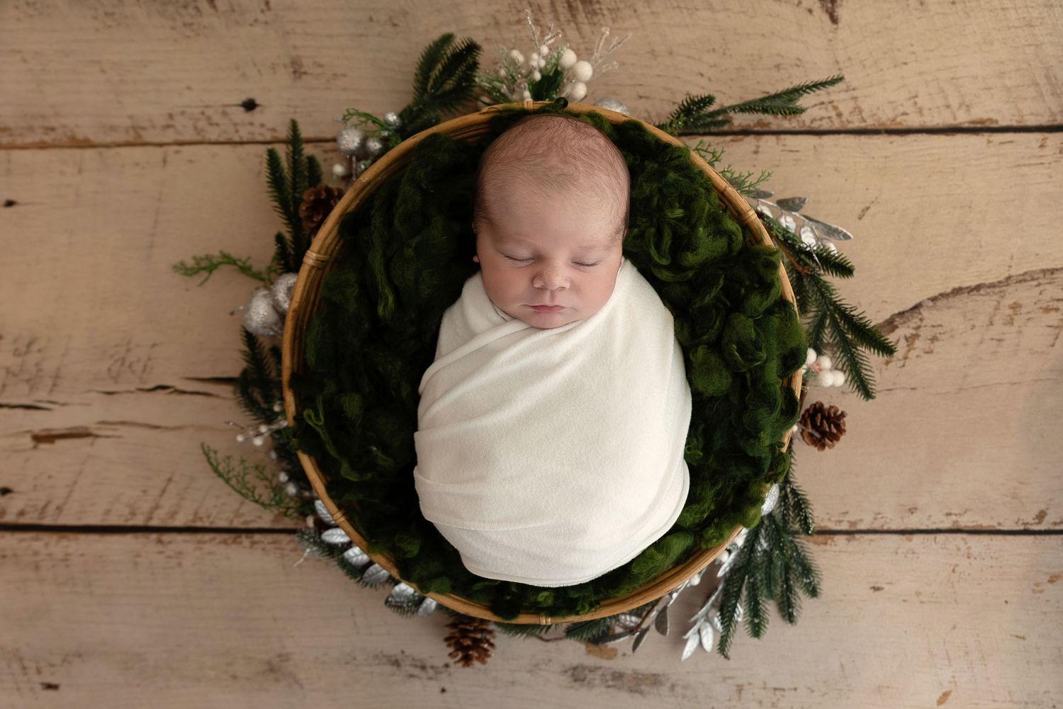 Sleeping newborn wrapped in white swaddle blanket nestled in round wreath with pine cones and greenery on wooden floor.