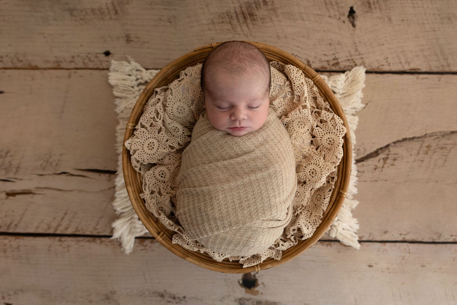 A sleeping newborn swaddled in beige wrap lies in a round wooden bowl with lace accents on rustic wooden floor.