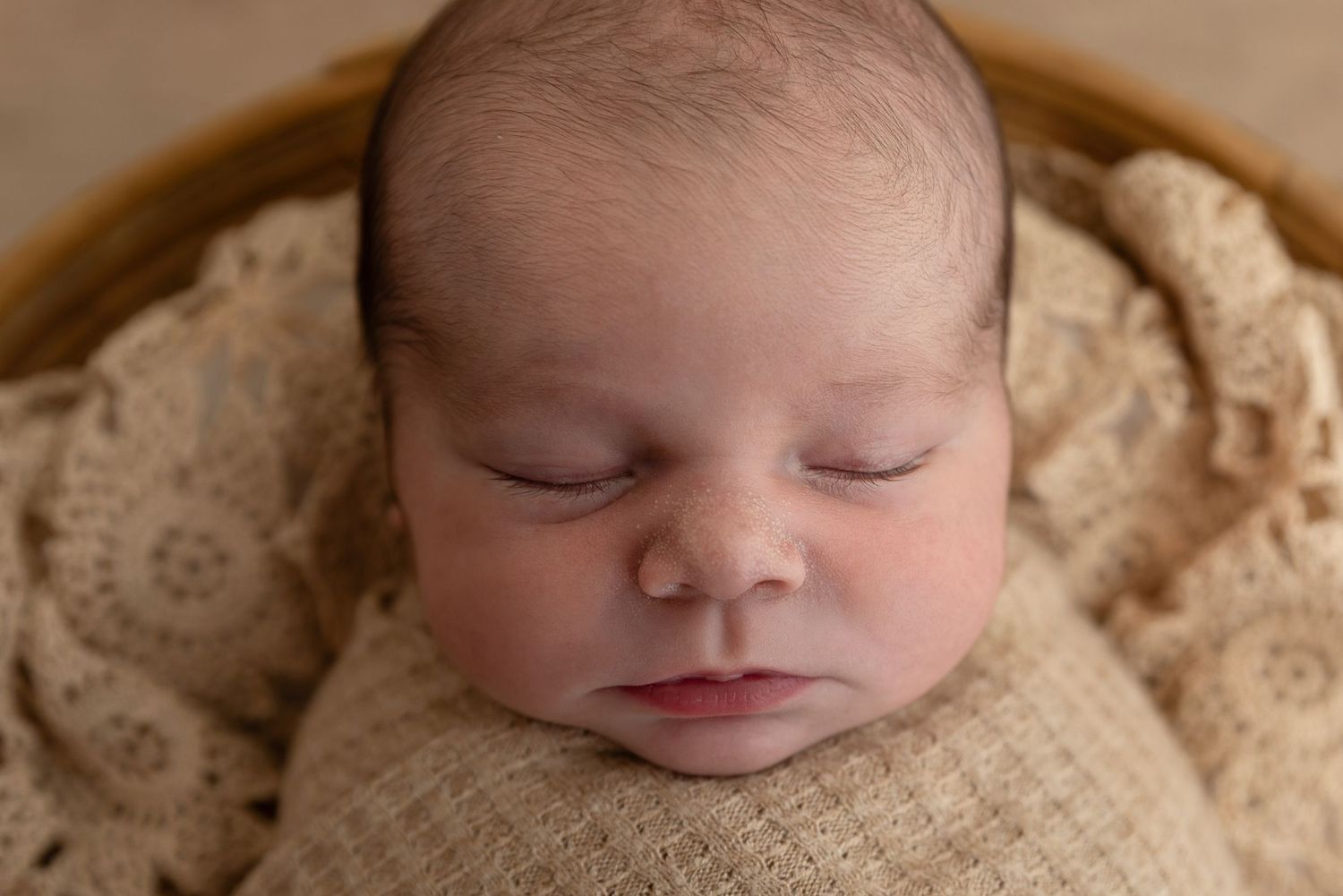 Peaceful sleeping newborn baby wrapped in soft beige knit blanket during professional portrait session.