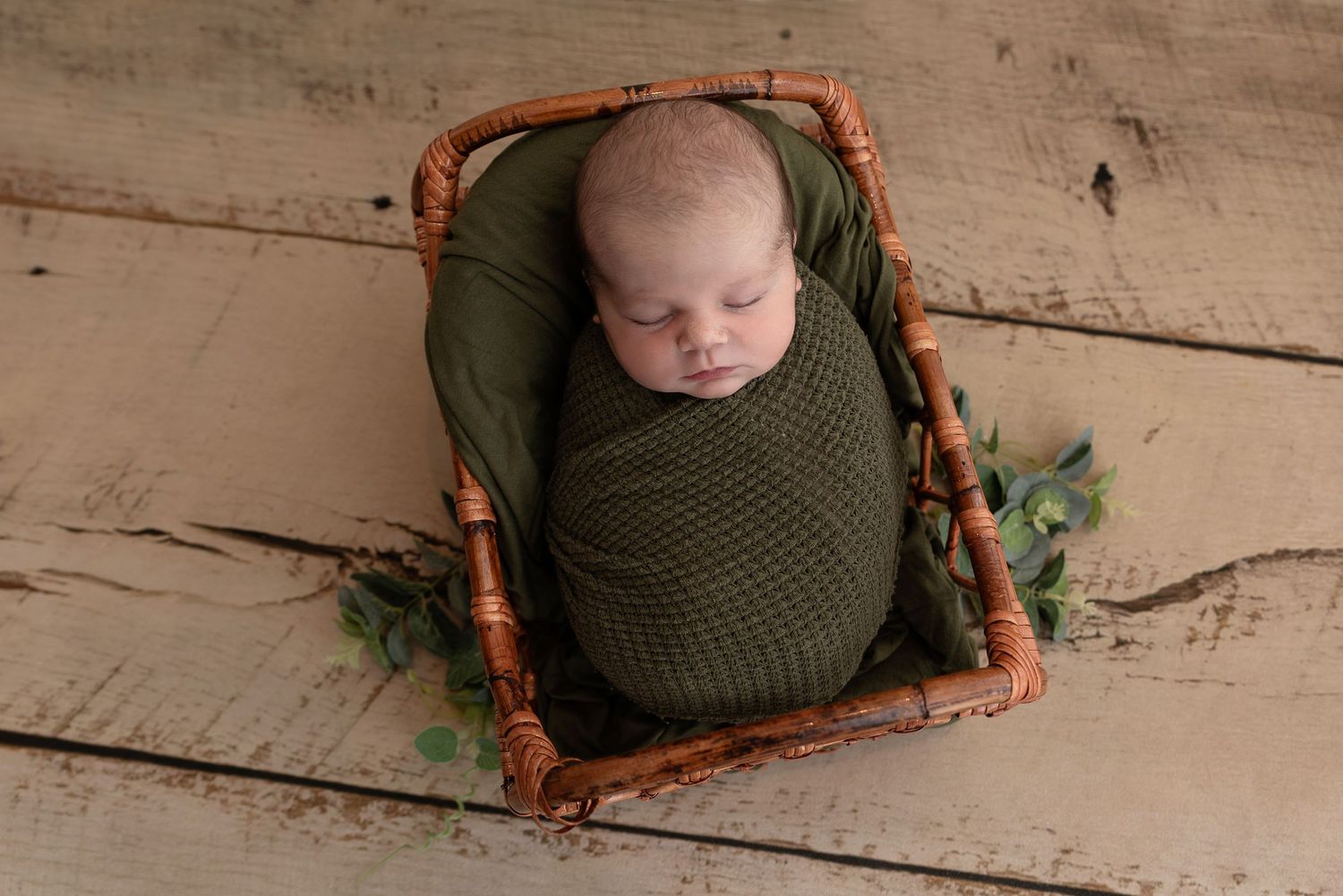 A sleeping newborn wrapped in olive green knit blanket rests in a wooden basket with greenery on wood floor.