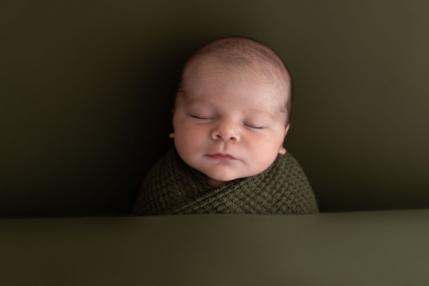 A sleeping newborn wrapped snugly in a brown knit blanket against a dark background.