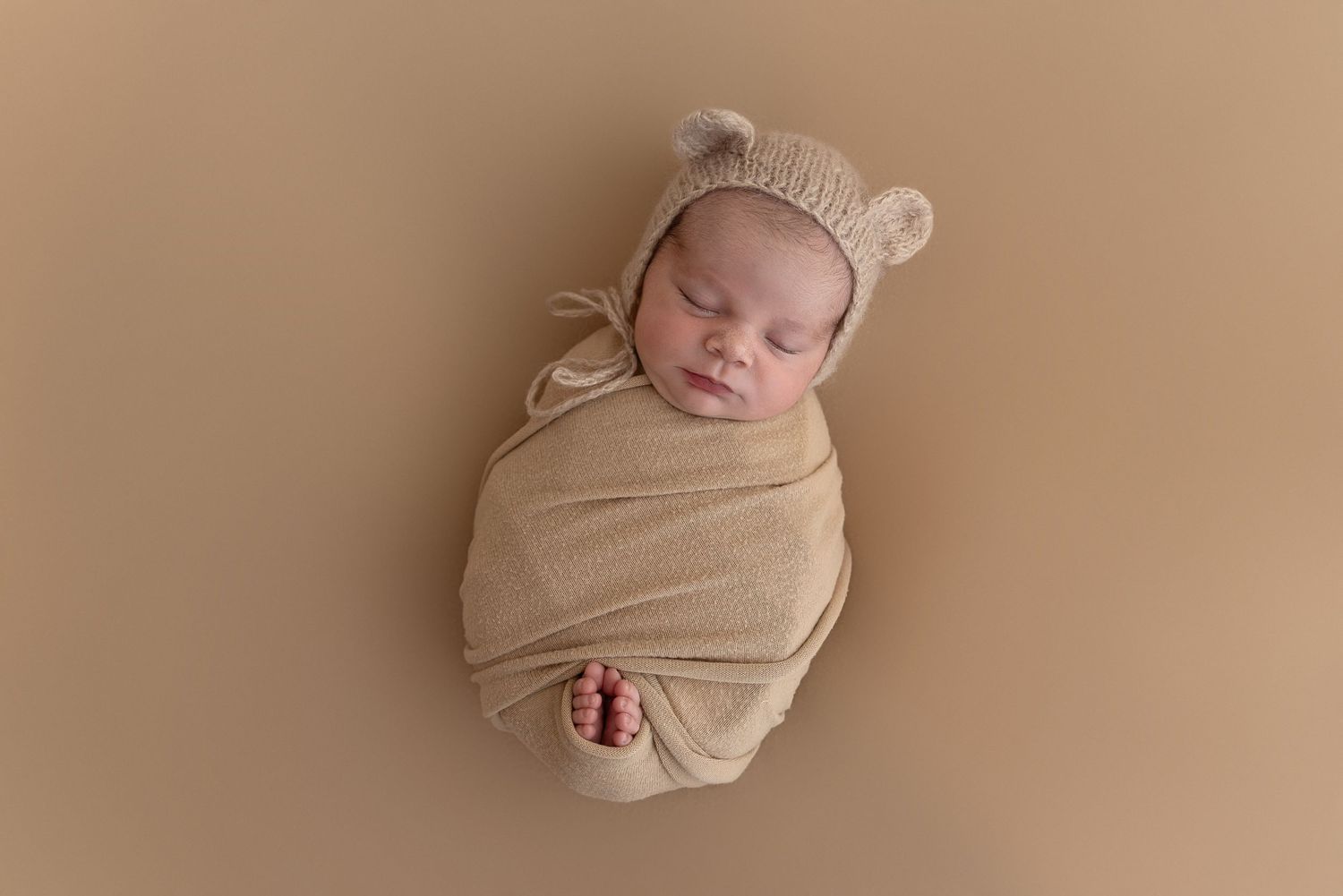 A peacefully sleeping newborn wrapped in beige swaddle wearing a knitted bear ear hat on brown backdrop.
