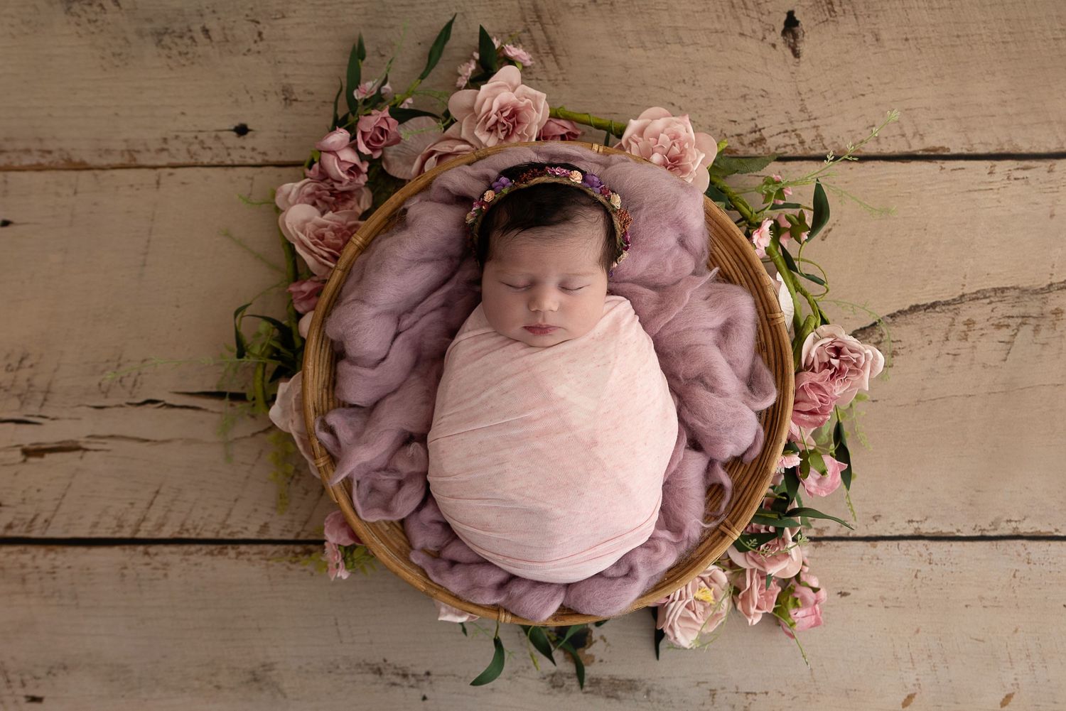 A swaddled newborn rests in a purple nest surrounded by pink roses on wooden floorboards.