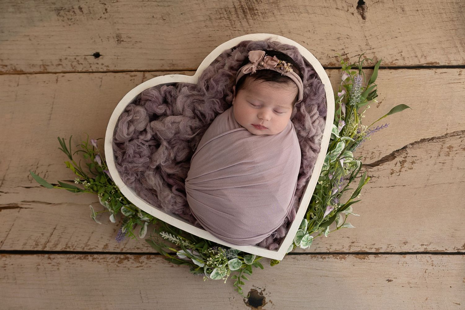 A sleeping newborn wrapped in mauve swaddle lies in a heart-shaped bowl with greenery on wooden backdrop.