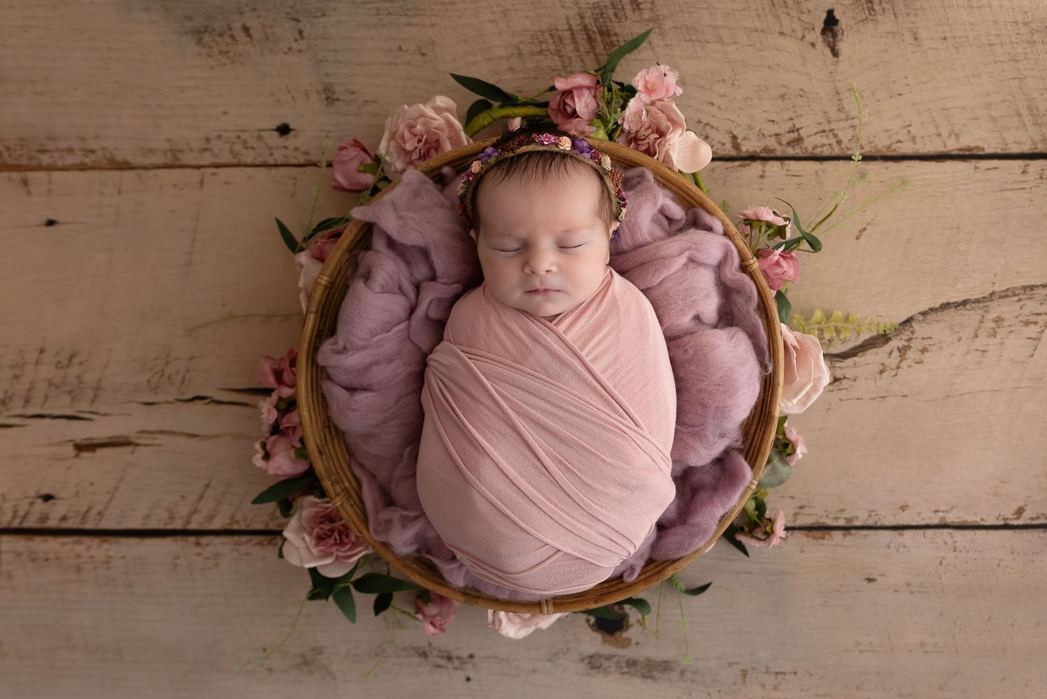 Sleeping newborn baby wrapped in pink swaddle in wooden bowl with flowers on rustic wooden background.