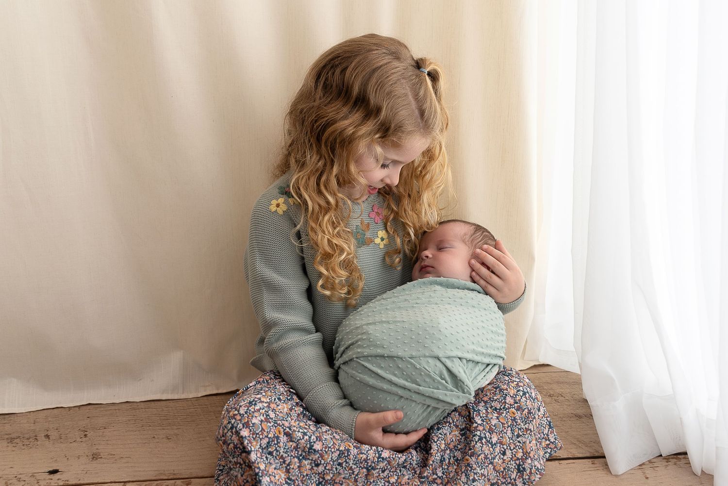A tender moment shows two people sitting by a window in soft natural light, wearing sage green and floral attire.